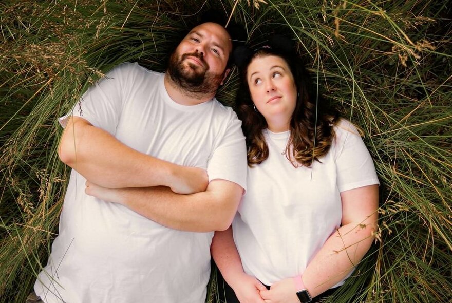 Two people lying on grass wearing white T-shirts, man with beard with arms folded, woman looking perplexed with hands clasped.