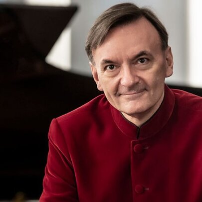 A portrait of pianist and composer, Sir Stephen Hough, wearing a red velvet button-up jacket, sitting in front of a grand piano.