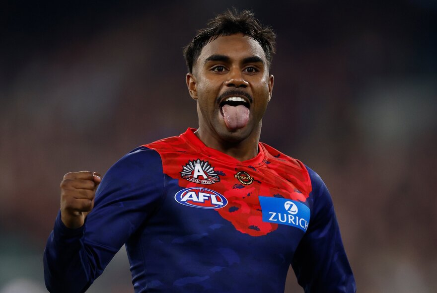 A Melbourne AFL player celebrating on the field with his tongue hanging out and his fist clenched.