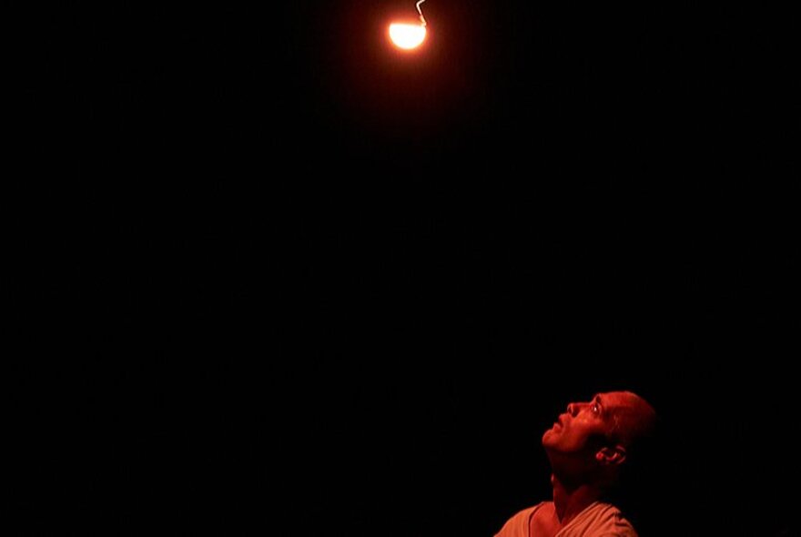 A single, warm light bulb shining from the top of a dark stage, with a person's head and shoulders in the bottom corner illuminated by a red glow as they look straight up at the light.