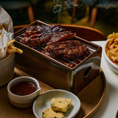 Bowls of chips and condiments next to two pieces of meat steaming on a griddle on a tray.