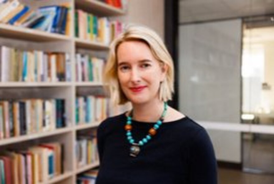 Author Jessica Lake, a woman, with blonde hair wearing a black top smiles in front of a bookshelf.