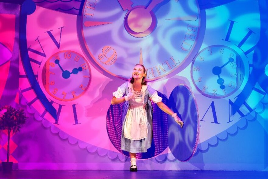 An actor in the role of Alice, performing in the family theatre show of Alice in Wonderland, stepping out of a giant clock prop on stage.