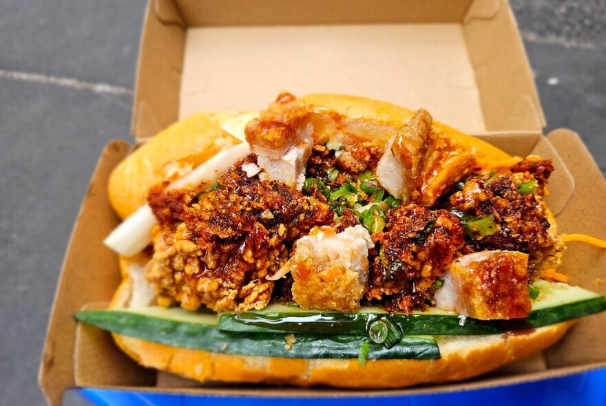 A cardboard container with a banh mi inside heaving with crispy pork chunks.