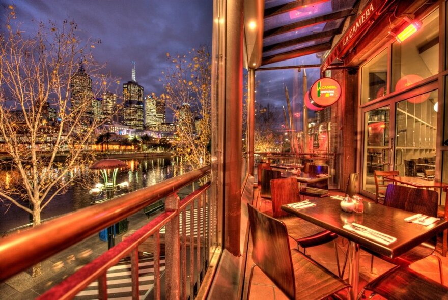 Balcony dining area at La Camera restaurant, overlooking the Yarra River and with city building views in the background, twilight.