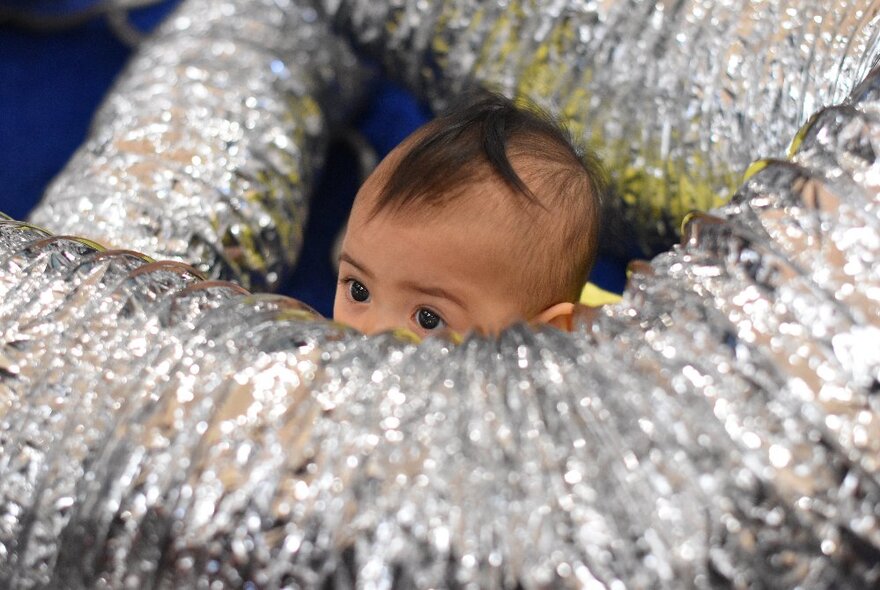 A baby peeking out of a tangle of silver flexible pipes. 