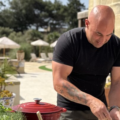 A chef in a black t-shirt, leaning over an outdoor prep bench in a sunny Italian locale, with a red enamel pot.