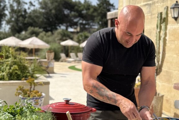 A chef in a black t-shirt, leaning over an outdoor prep bench in a sunny Italian locale, with a red enamel pot.