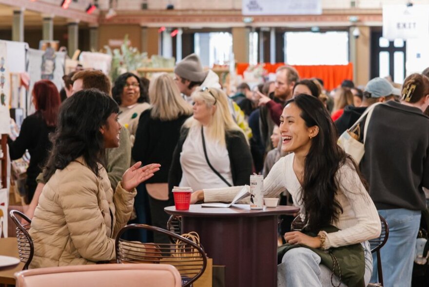 Two friends having refreshments and interacting at a table and chairs at an indoor design market, with crowds of people wandering around the exhibition space behind them.