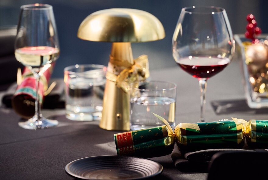 A festive table setting at a restaurant featuring a small brass lamp, wine glasses, water tumblers, and a Christmas cracker.