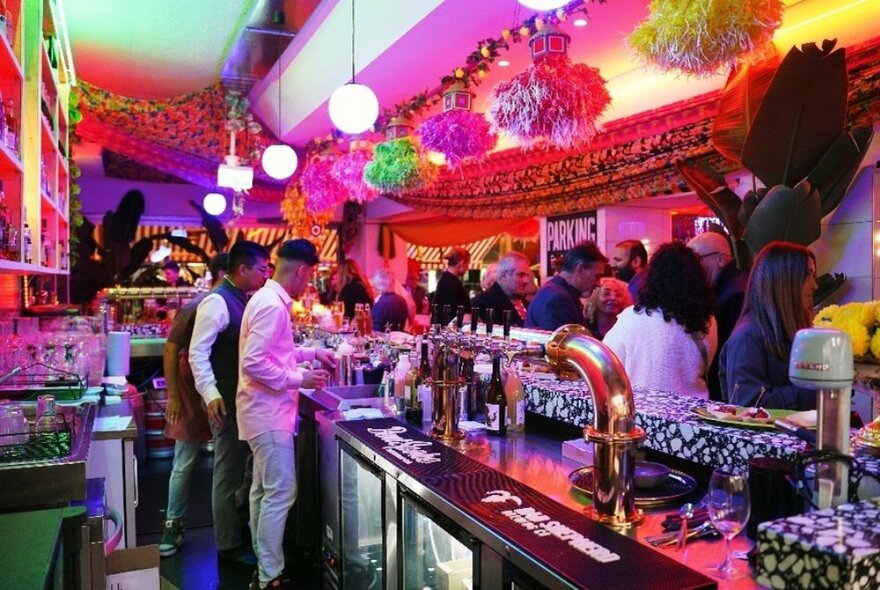 A brightly decorated bar from behind the counter with staff working and patrons mingling under large paper pop-poms.