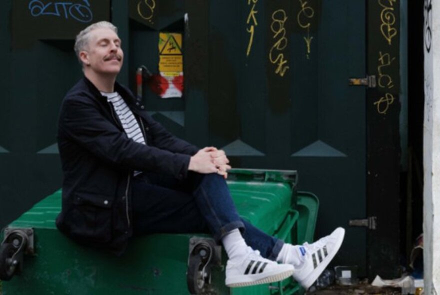 Comedian Carl Donnelly, sitting back on a large green case and smiling. 