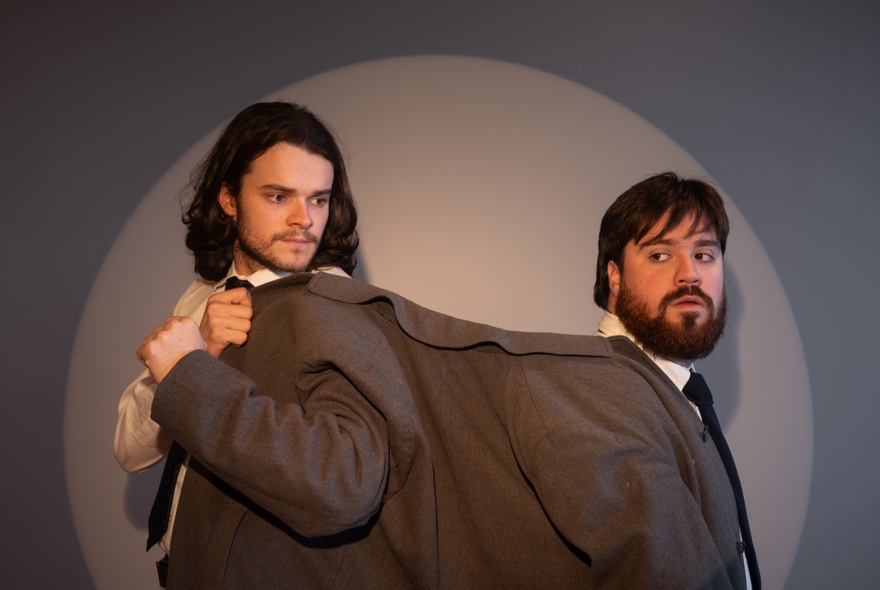 Two men in a spotlight standing back to back, each wearing one arm of a brown jacket stretched between them.