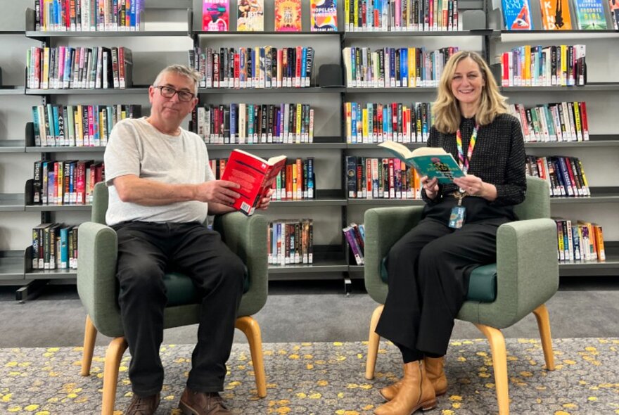 Two people seated in armchairs in front of library shelves, each holding books and smiling.