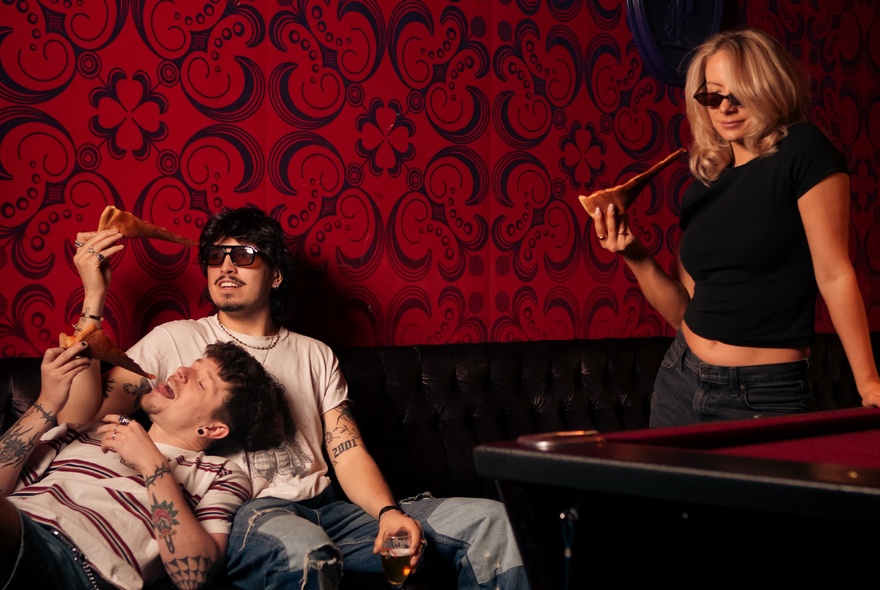 Three people eating slices of New York-style pizza in a room with dark red wallpaper and a pool table.