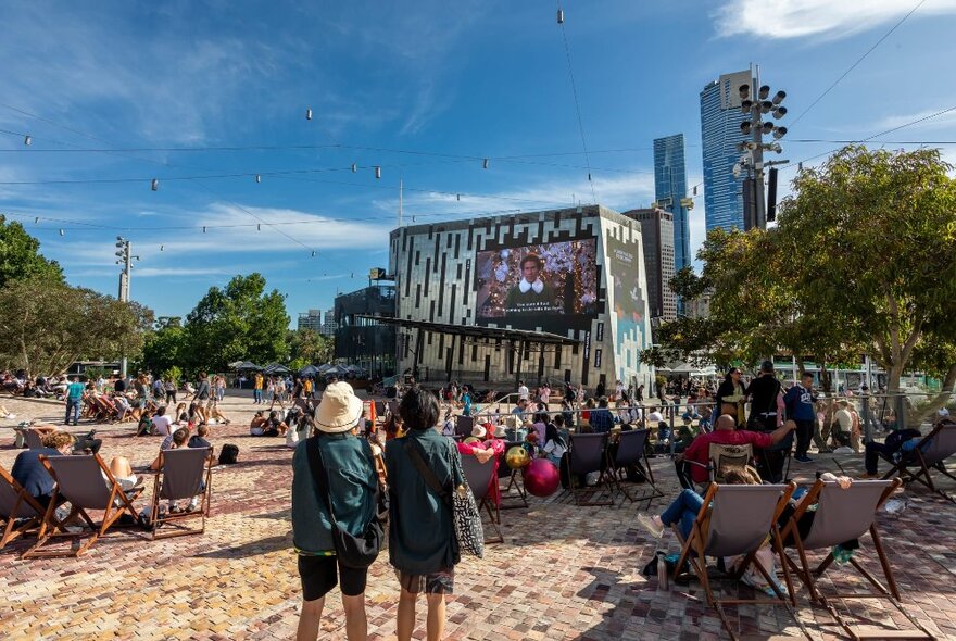 People gathered in the main plaza at Fed Square, seated on deck chairs or rugs, facing towards the big screen and watching a Christmas movie.