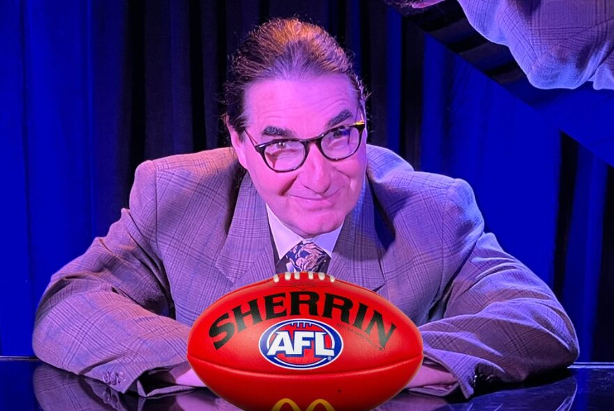 A man in a grey suit and glasses, seated at a shiny table with a bright red Sherrin AFL football in front of him.