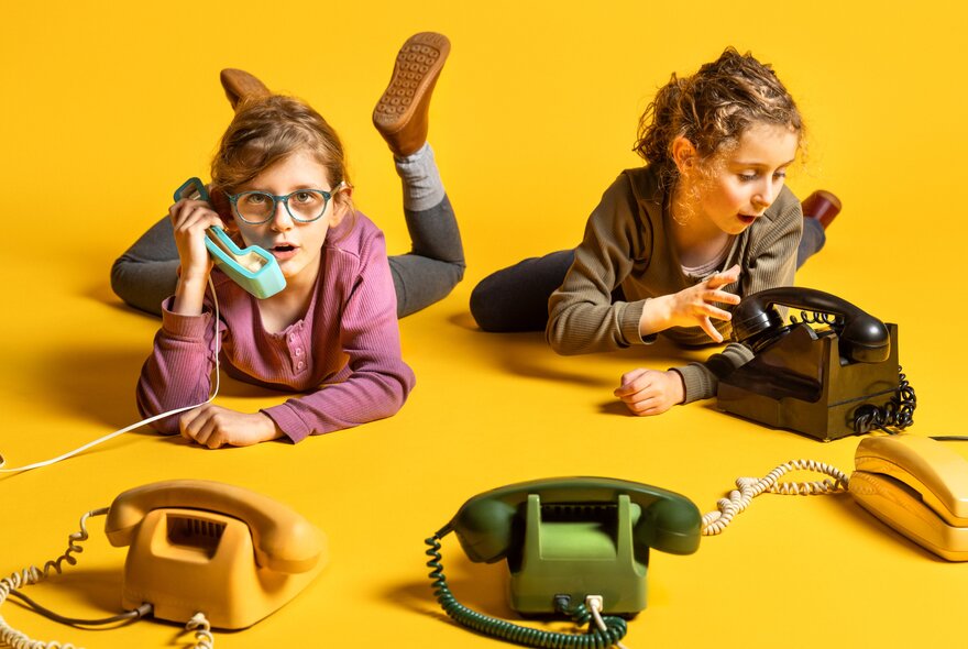 Two girls on their tummies on a yellow floor talking on landline telephones. 