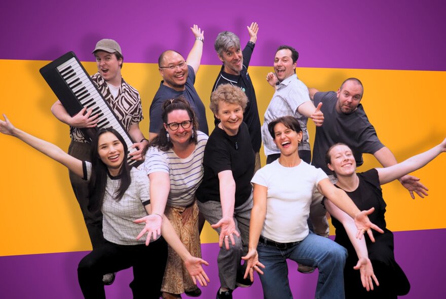 A cast of actors posing happily for a photo, with arms outstretched, against a purple and yellow background.