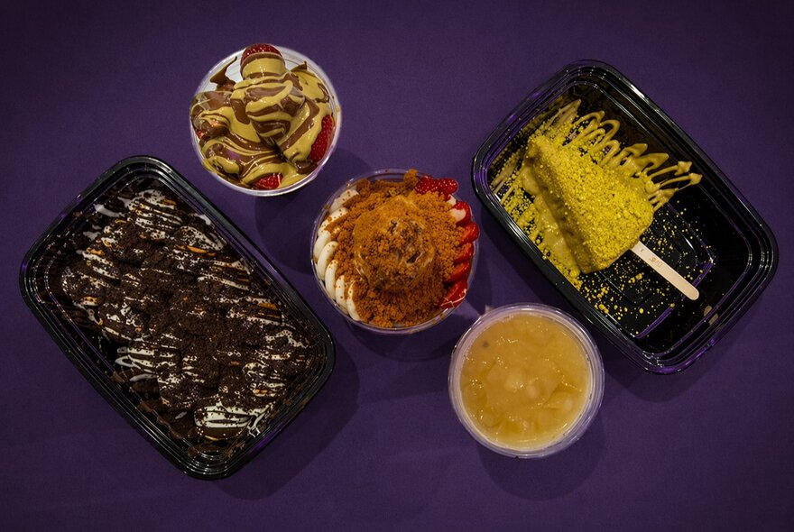 Top down view of a selection of sweet treats in take-away containers on a purple table top.