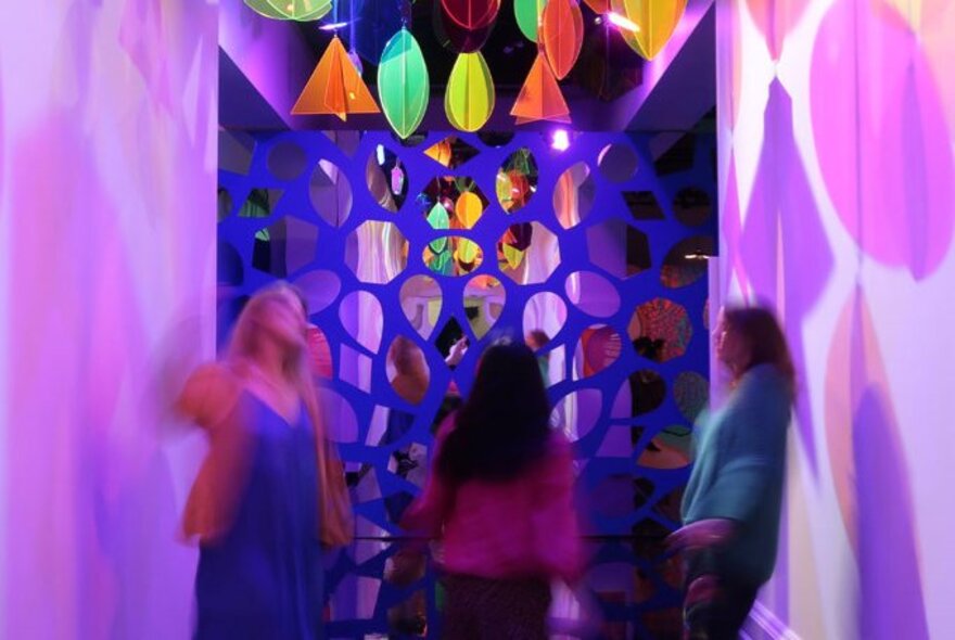 Three people blurred as if in motion inside a colourful room at an exhibition inside the Immigration Museum.