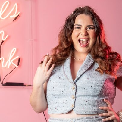 Cabaret artist and vocal coach Amelie Peters posing in front of a pink neon wall sign that reads Pals in Pink, against a solid pink background.
