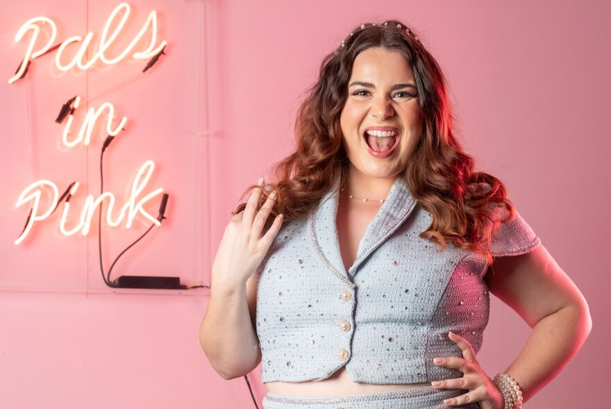 Cabaret artist and vocal coach Amelie Peters posing in front of a pink neon wall sign that reads Pals in Pink, against a solid pink background.