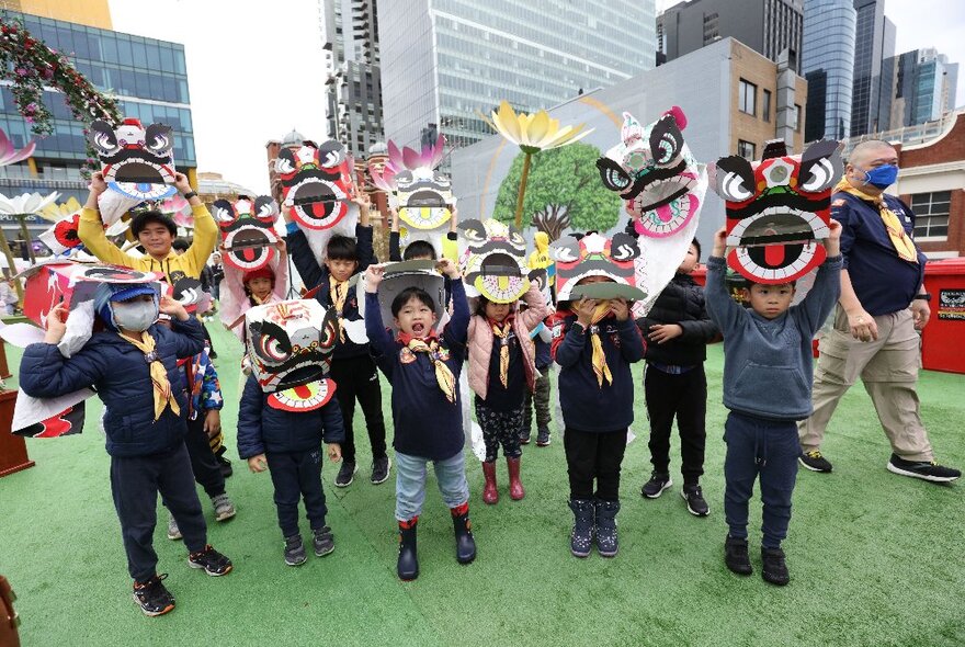 Group of children holding Chinese lion dance masks on an astrotuf stage surrounded by city buildings.