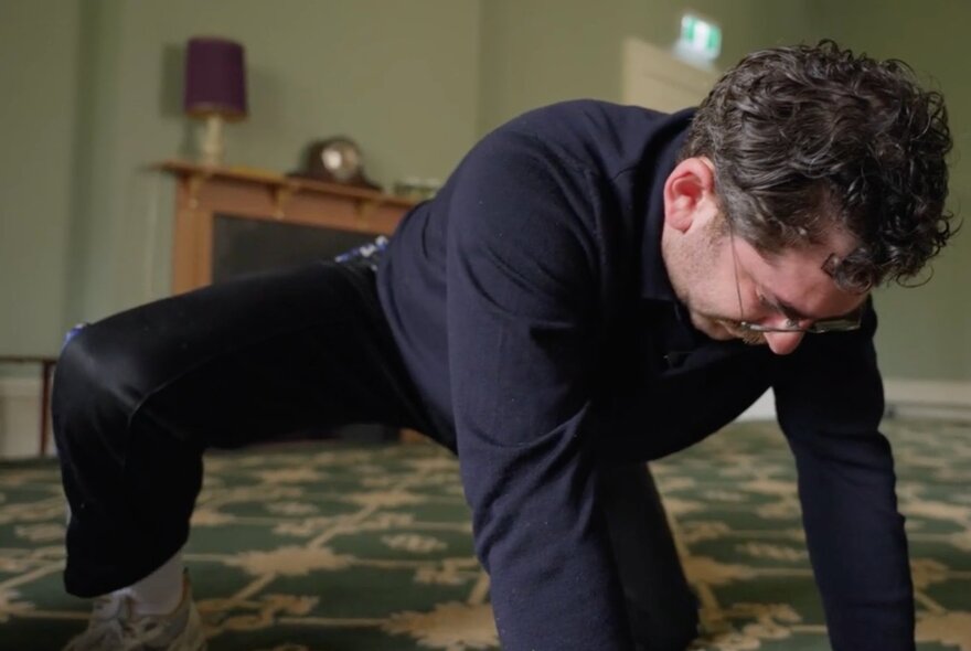 A man with glasses and dark hair crouching on the floor of a carpeted room, appearing to be in a state of deep concentration or physical exertion.