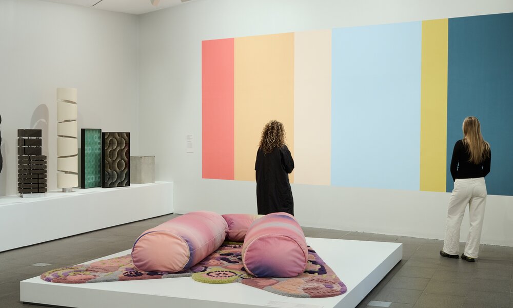 Two women are in a gallery looking at a colourful striped painting. There is a large pink soft sculpture behind them in the shape of a giant U.