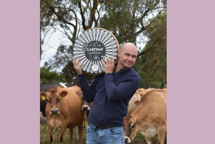 A man holding up a huge wheel of cheese, outdoors with cows behind him.