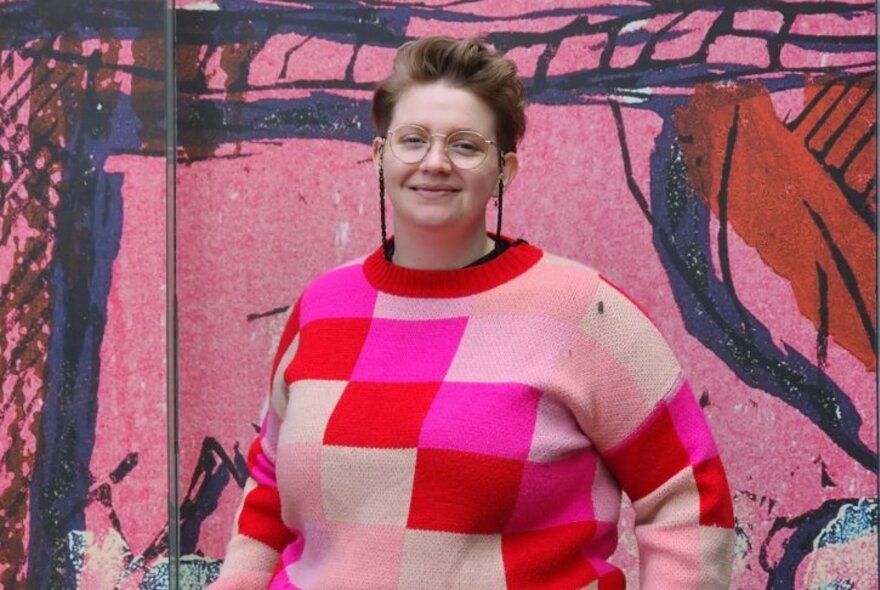 A portrait of writer, Sarah Giles, in a pink checked jumper, standing in front of a pink painted mural.