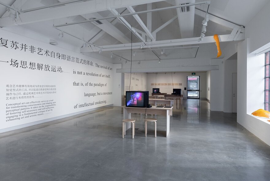 A large white space in a gallery, with text written directly on the wall, and a small table in the centre of the room with a monitor on it.