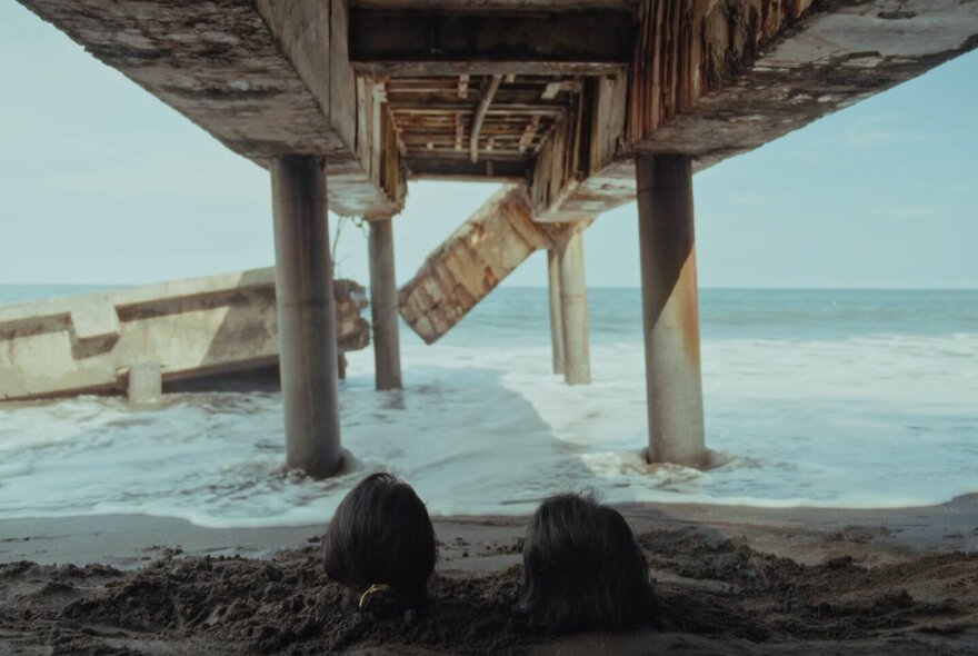 A pier extending into the sea from the land, part of it broken and half submerged in the shallow water, in the foreground two human  head are poking out from the sand and facing the horizon.