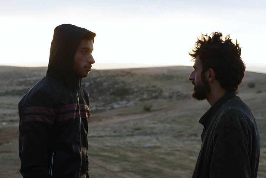 Still from the documentary film No Other Land showing two men in profile talking to each other, a large field in the background.
