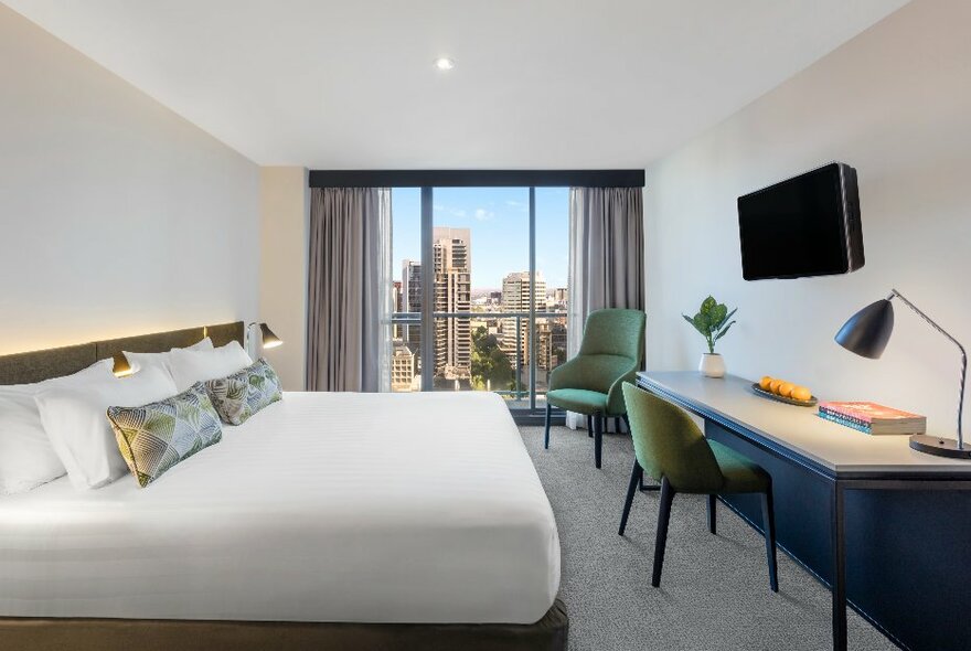 Hotel room with white bed, long desk against one wall and curtains framing a window looking onto city views.