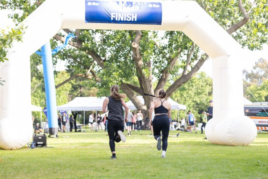 Two people running through the inflatable arched finish marker of a fun run in a park.