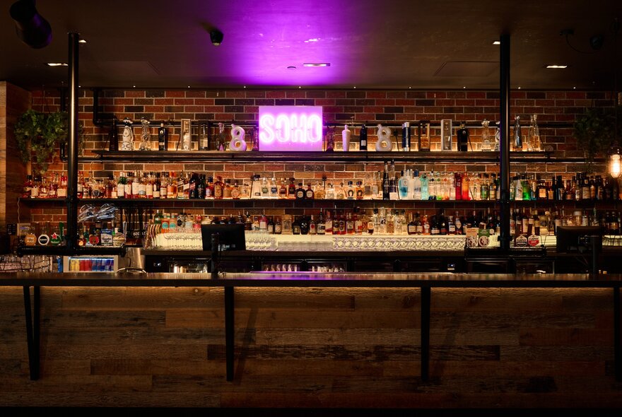 An interior of a dimly lit bar with a rustic, industrial vibe, the rear wall filled with bottle on shelves.