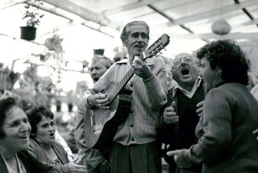 Italian men and women singing in a light- and plant-filled outdoor space, one man playing the guitar.