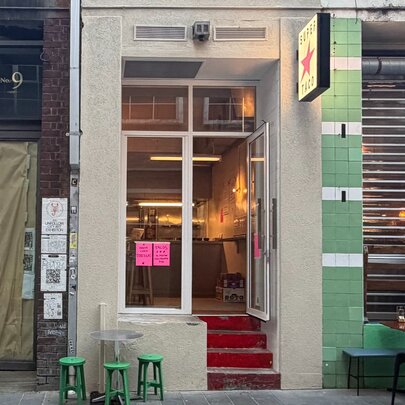 A very narrow shopfront with four stairs going up to the glass door, sandwiched between two other very narrow shopfronts.