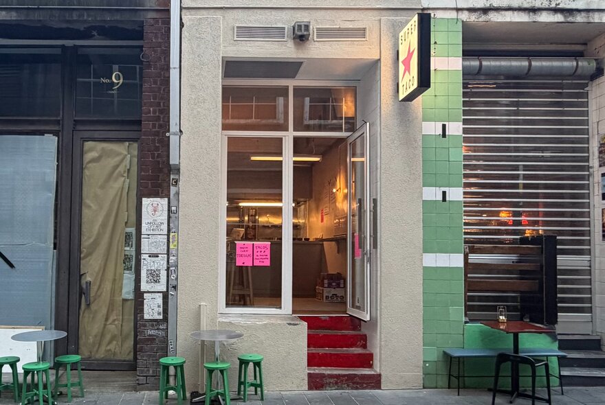 A very narrow shopfront with four stairs going up to the glass door, sandwiched between two other very narrow shopfronts.