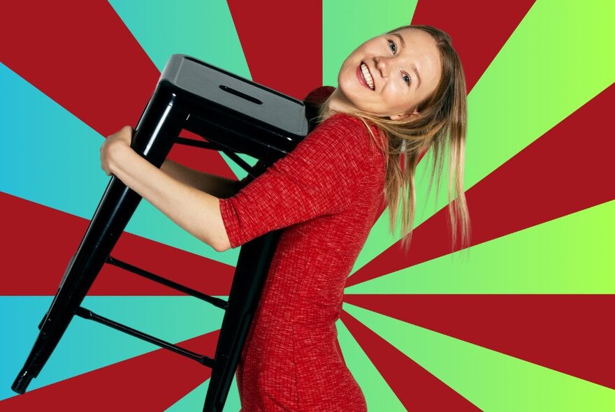 A young woman clutching a black kitchen stool to her chest in an embrace with a sunburst pattern of red and blue/green stripes behind her. 