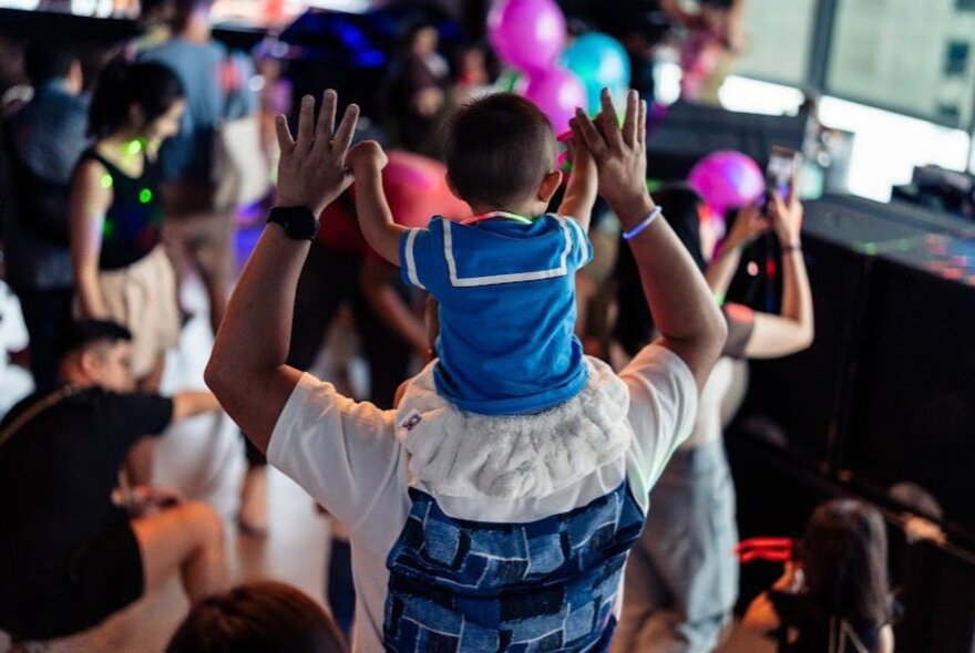 A baby on her Dad's shoulders at a kid-friendly dance party; other kids and parents in the background.