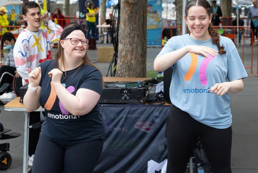 Two people smiling and dancing in front of a DJ's mixing desk, at an outdoor festival.