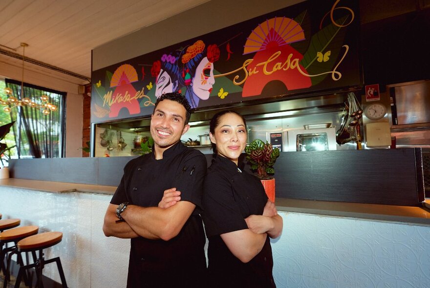 A pair of chefs wearing black, standing back to back in front of the bar at a restaurant.