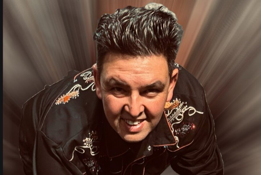 A man with big hair crouching down and looking straight ahead with dark coloured beams all around him. 