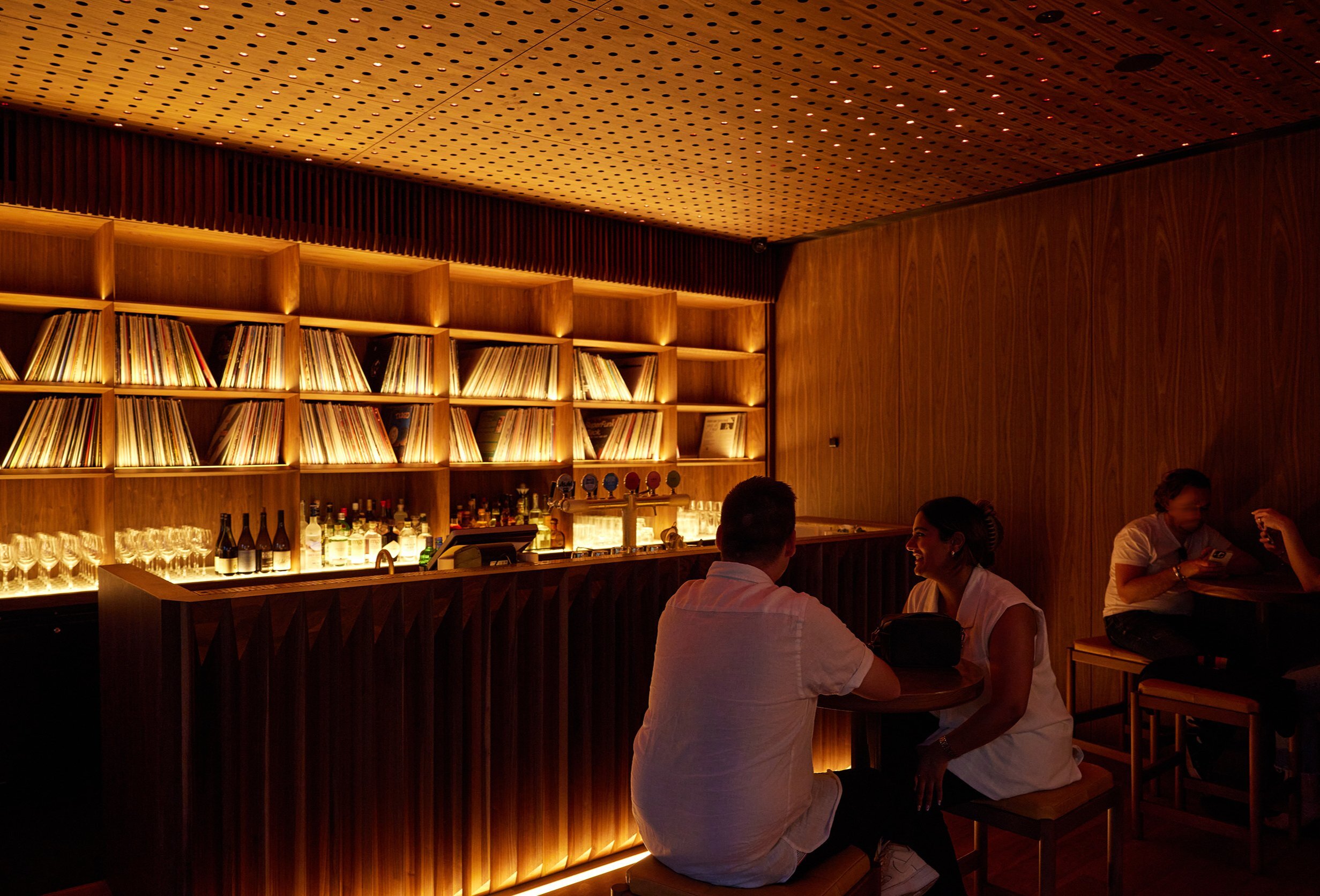 Two friends are in a dimly-lit bar surrounded by vinyl records.
