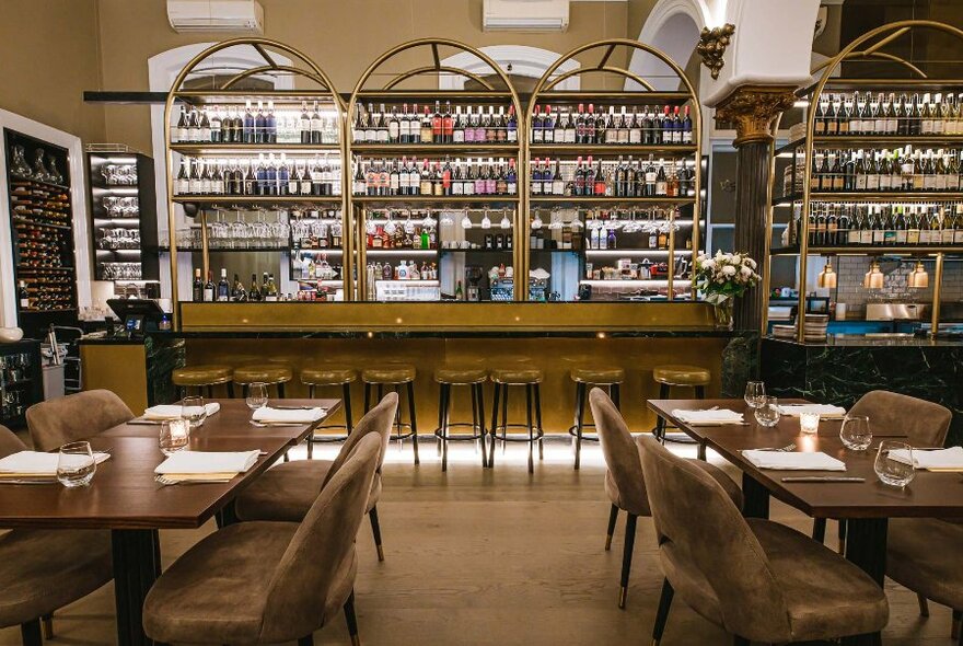 Interior of restaurant with tables and chairs set for service and a view onto a rear bar, which is flanked by shelves of bottles of wine.