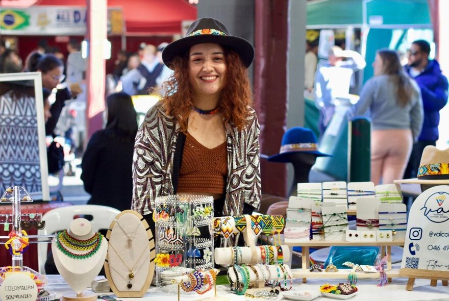 A woman in a hat standing behind a market stall with displays of hand-made jewellery.