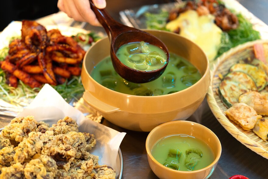 A person scooping up matcha Korean rice wine from a large yellow bowl surrounded by an assortment of Korean dishes.